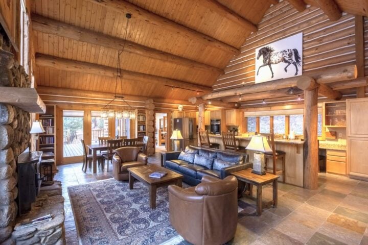 The spacious log cabin interior at Triple Creek Ranch boasts wooden beams, a stone fireplace, leather furniture, and a patterned rug. A horse painting enhances the warm atmosphere, while large windows and a dining area complete the cozy setting.