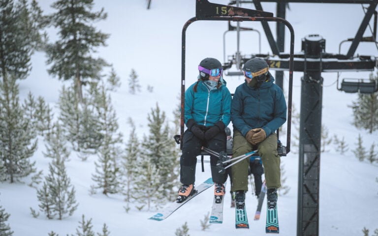 Two people sitting on a snowboard on the Lost Trail Ski Area ski lift in the snow.