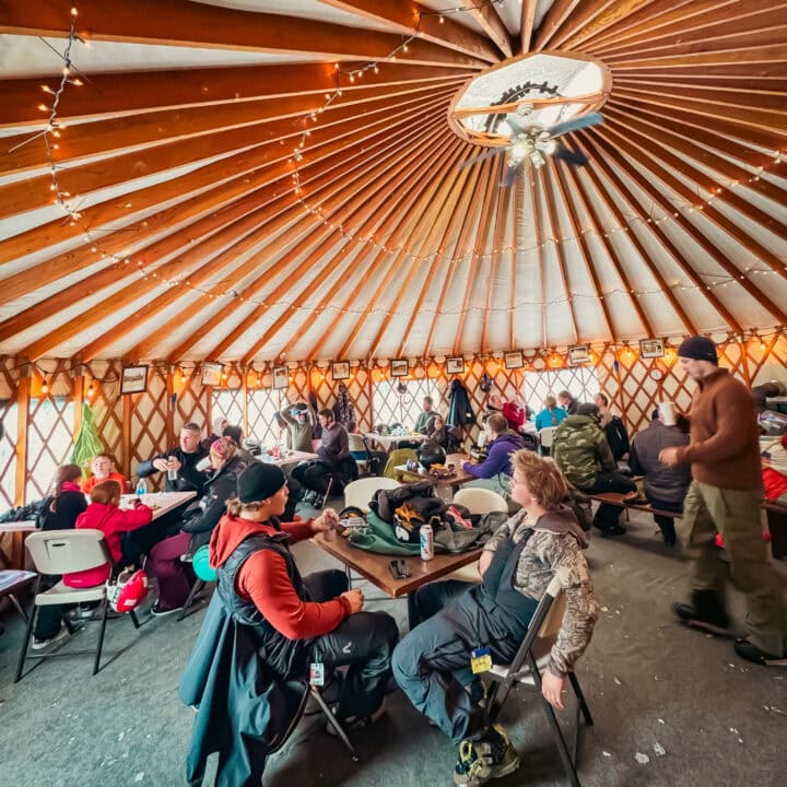 A group of people in winter clothing gather inside a yurt with a wooden roof and string lights, engaging in conversation and activities. The atmosphere feels as cozy as an Elementor-designed space, blending style and comfort seamlessly.