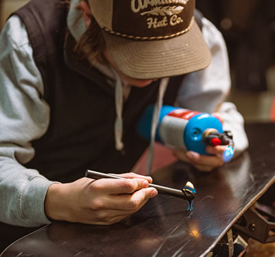 A person in a cap uses a blowtorch to drip liquid onto a black surface, crafting what could be the perfect header design with unparalleled precision.