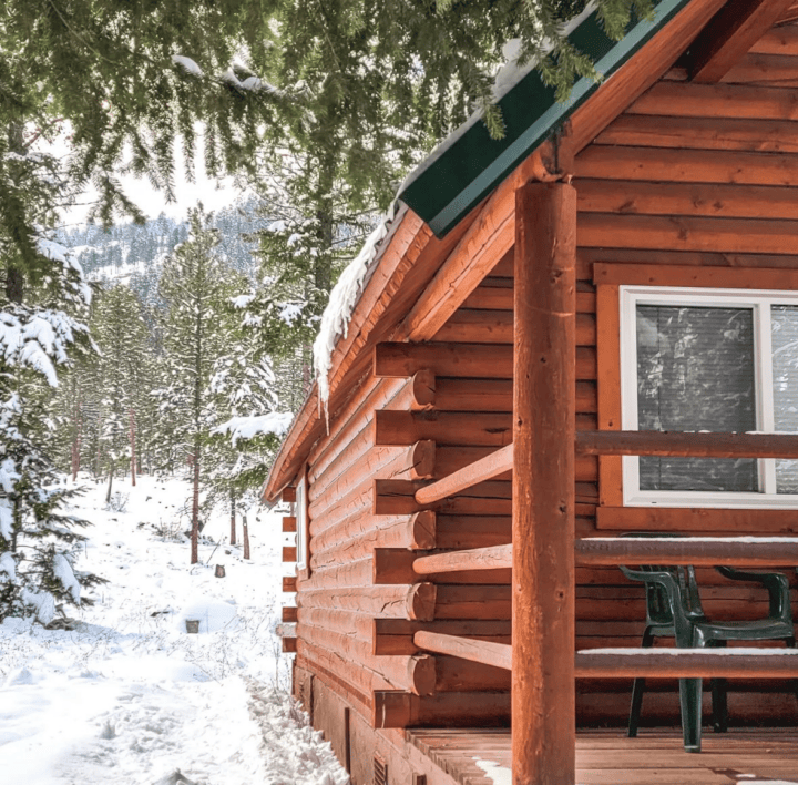 The wooden cabin, a cozy lodging option, features a covered porch surrounded by snow and trees. A plastic chair rests on the porch, inviting you to take in the serene view of the snow-covered mountains visible in the background.