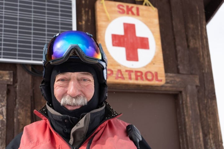 An older man in ski gear stands in front of a ski patrol building with a red cross sign at Lost Trail, highlighting essential ski patrol jobs on the mountain.
