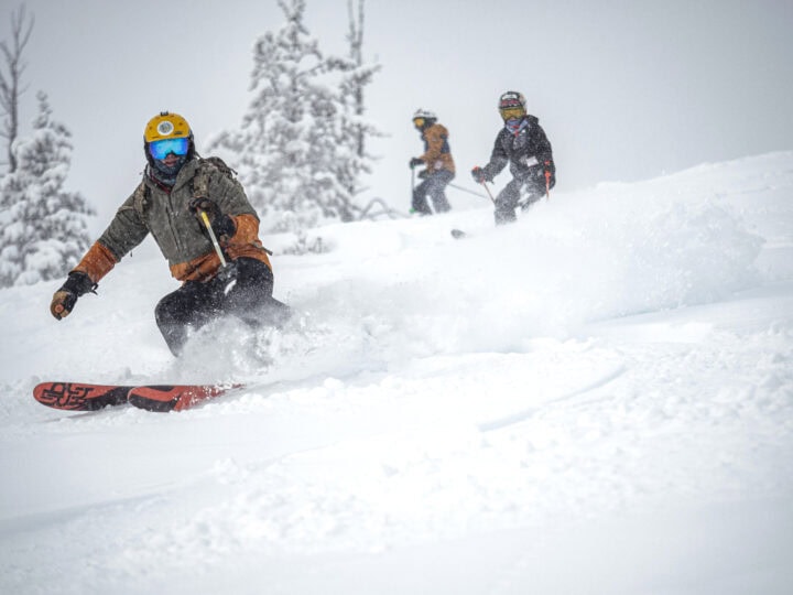 Three people are snowboarding and skiing down a snow-covered slope at Powder Mountain. The foreground shows a snowboarder in motion, while two skiers follow in the background. Trees covered with snow are visible.