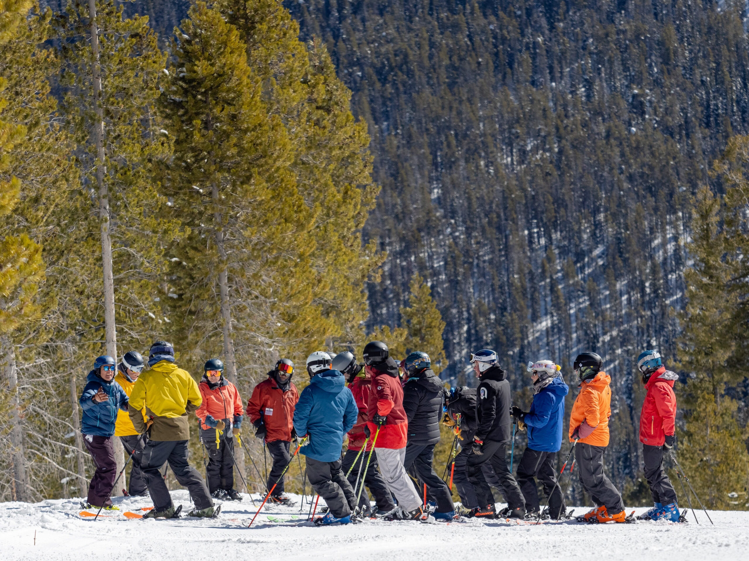 A group of people in colorful ski gear gathers on a snowy slope at Lost Trail, surrounded by towering pine trees, capturing the thrill that Deb Armstrong once epitomized.