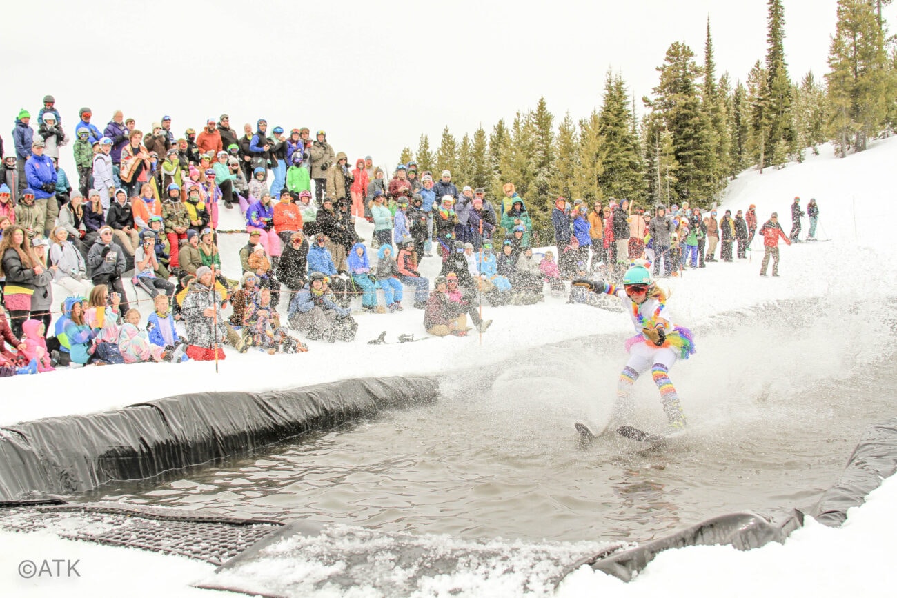 A skier in colorful clothing attempts to cross a snowy pond while a large crowd watches from the side at a winter event.