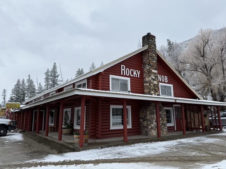 A red log cabin building labeled "Rocky Knob," with a stone chimney, sits among snow, trees, and parked vehicles—a welcoming spot for visitors and local partners alike.